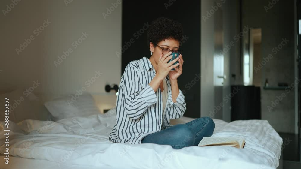 Happy Hispanic curly haired woman reading a book and drinking tea on the bed at home Happy Hispanic curly haired woman reading a book and drinking tea on the bed at home