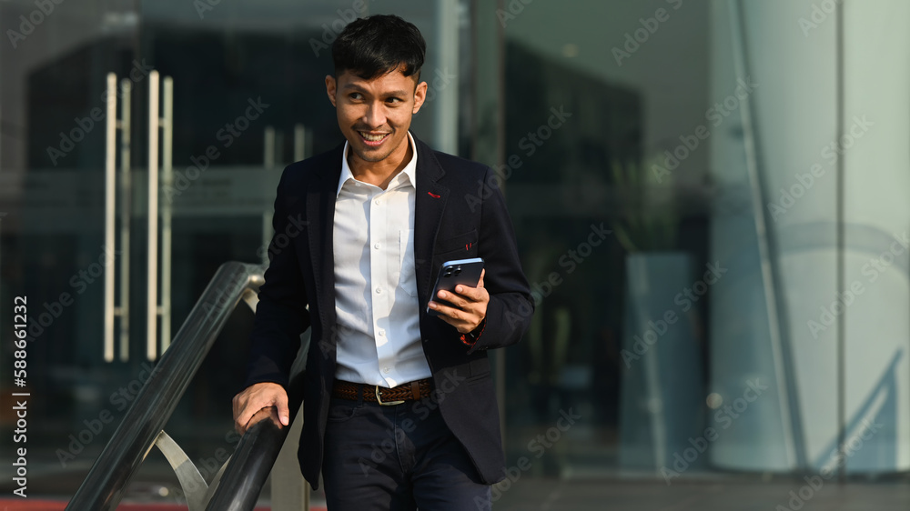 Naklejka premium Smiling young businessman holding smart phone walking up stairs and looking away. Urban lifestyle and technology concept