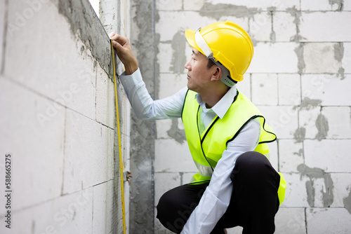 Young Asian engineer in engineering uniform and helmet at construction site using a measurement tape to measure for precision. 30s professional foreman working on structure building at workplace