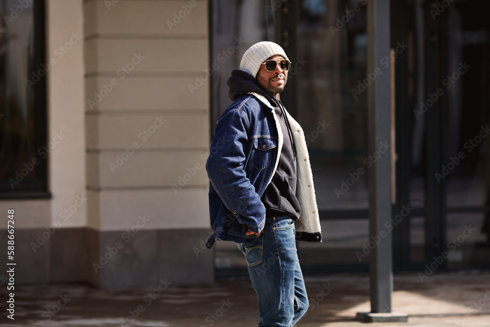 Handsome African american man model wearing stylish outfit denim jacket, hat, hoodie and sunglasses walking in city Street