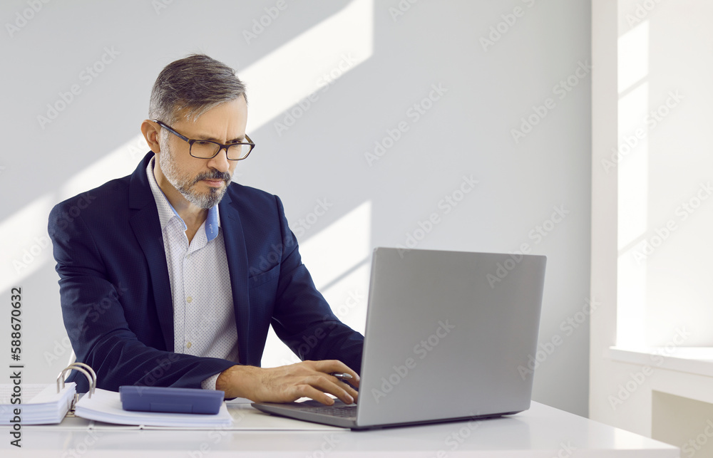 Serious concentrated businessman at workplace is engaged in calculating finances of his company. Man in office sitting at table with calculator and paper documents and looking at laptop screen. Serious concentrated businessman at workplace is engaged in calculating finances of his company. Man in office sitting at table with calculator and paper documents and looking at laptop screen.