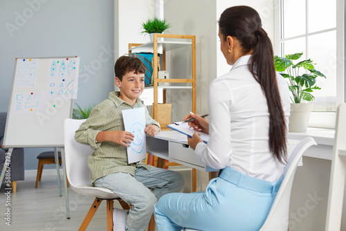 Cheerful relaxed preteen schoolboy talks about positive emotions with school psychologist. Child shows sheet with positive emoticon to female psychologist who is taking notes on clipboard. photo
