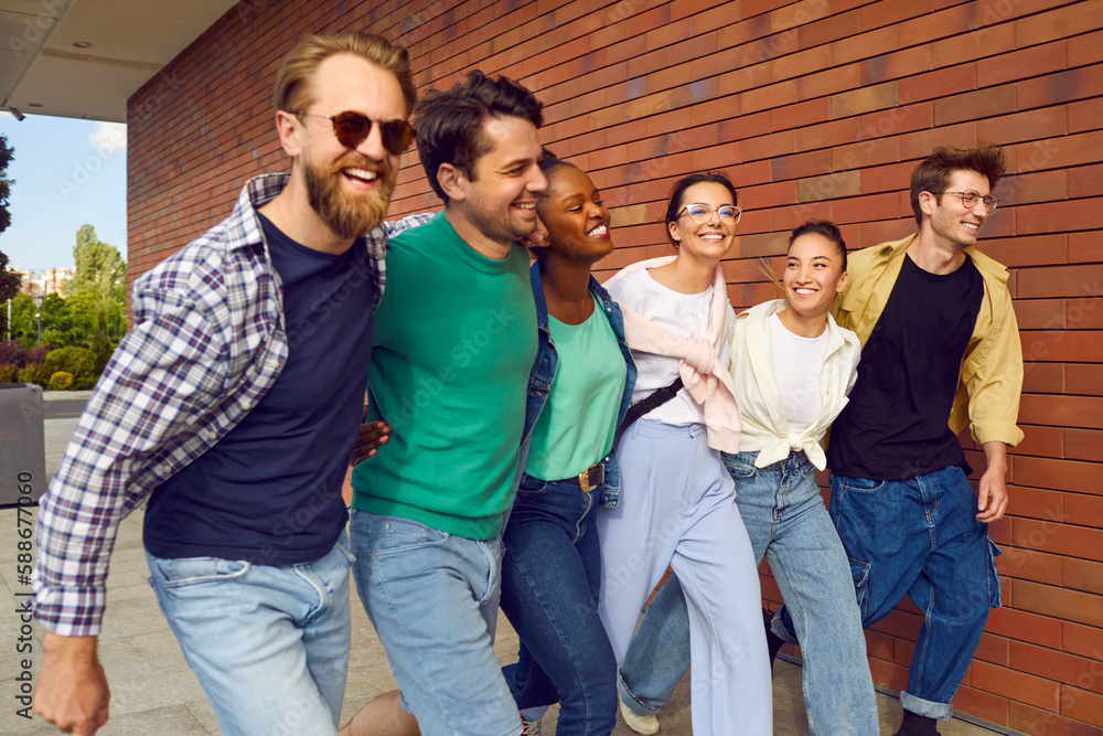 Meetings in city. Cheerful carefree group of friends in casual clothes are happily walking down street. Stylish multiracial young men and women smiling as they walk past brick wall. Youth concept.