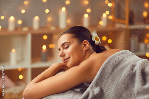 Young lady getting beauty treatments at a modern spa salon. Happy, relaxed woman with closed eyes lying on the bed, with beautiful candle lights gleaming in the background. Spa, female beauty concept