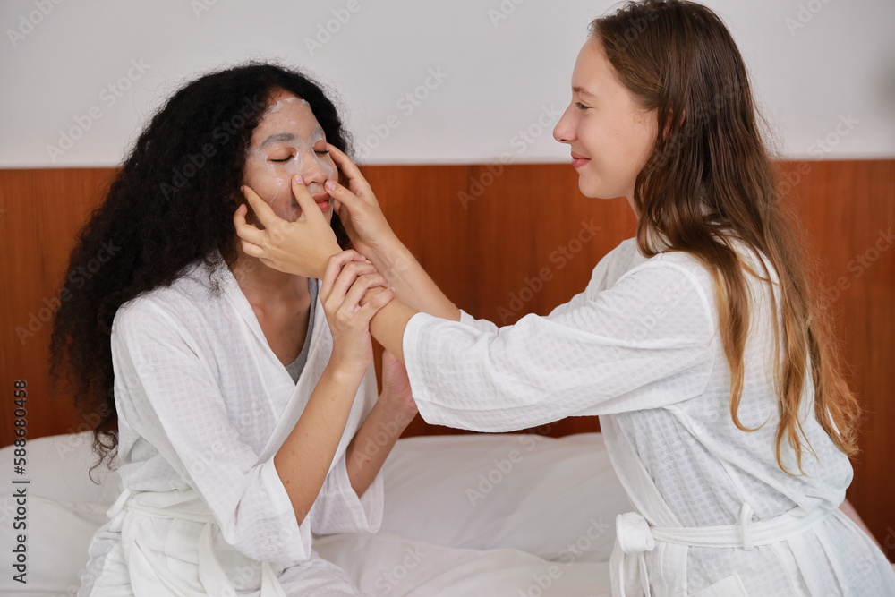 Young happy diversity ethnic beautiful woman in bathrobe sit on bed and doing cosmetic white facial mask for skin care and treatment. Women lesbian couple daily LGBTQ lifestyle and love concept. Young happy diversity ethnic beautiful woman in bathrobe sit on bed and doing cosmetic white facial mask for skin care and treatment. Women lesbian couple daily LGBTQ lifestyle and love concept.