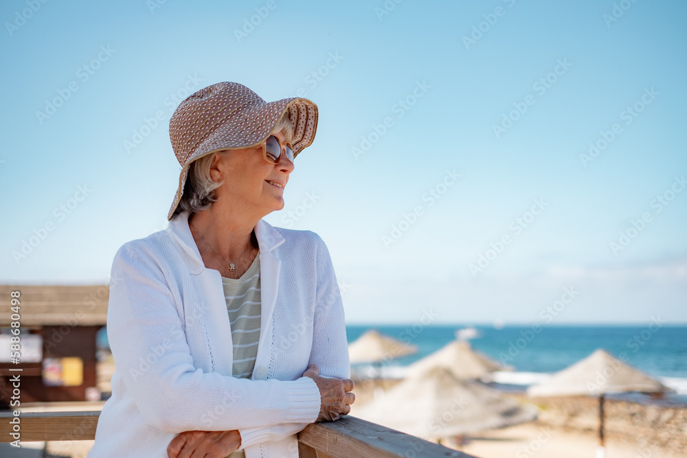 Smiling senior woman in casual clothing with hat and sunglasses relaxed at the beach looking away enjoying vacations and freedom during retirement - horizon over sea