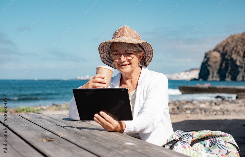 Smiling senior woman with hat sitting at wooden table at the beach enjoying reading pleasure holding digital tablet in hand, retiree lifestyle concept - horizon over sea and blue sky