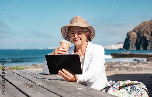 Smiling senior woman with hat sitting at wooden table at the beach enjoying reading pleasure holding digital tablet in hand, retiree lifestyle concept - horizon over sea and blue sky