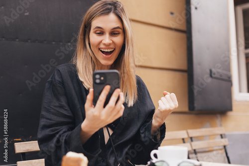 Joyful blonde woman playing video game on mobile phone, reading good news, celebrating winning lottery, feeling so happy and cheerful while making winner gesture, sitting at table with coffee cup.