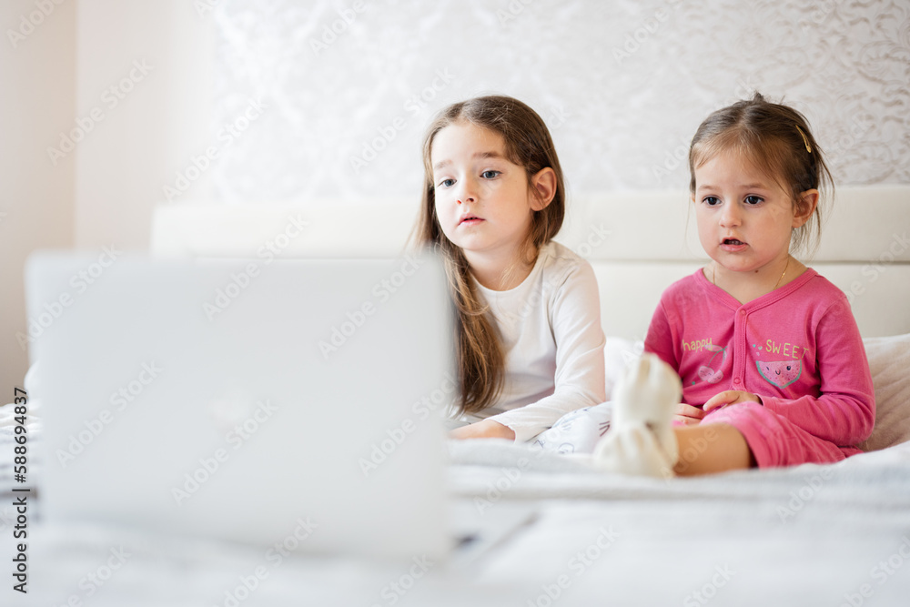 Two cute little sisters wear pajamas looking cartoon by laptop in morning at home. Two cute little sisters wear pajamas looking cartoon by laptop in morning at home.