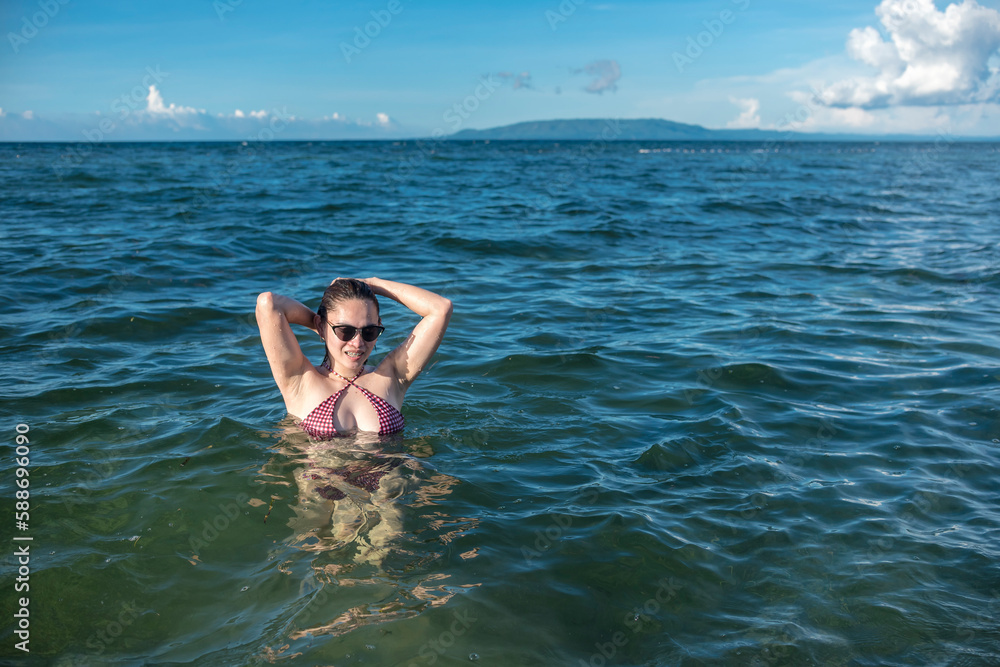 A sexy asian woman in a bikini and wearing shades wading in chest deep waters during a sunny day at the beach