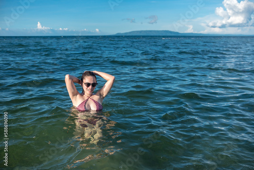 A sexy asian woman in a bikini and wearing shades wading in chest deep waters during a sunny day at the beach