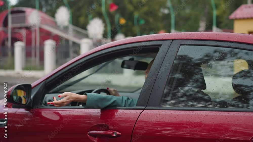 Covered with raindrops red car moves past amusement park. Positive female driver stretches hand through open window enjoying breeze while driving on blurred background Covered with raindrops red car moves past amusement park. Positive female driver stretches hand through open window enjoying breeze while driving on blurred background