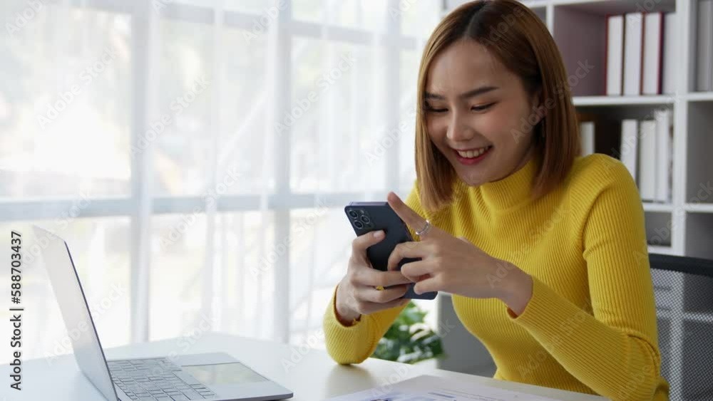 Young Asian women using a mobile phone while working at home. Female browsing sites, messaging, or shopping with a smartphone during a video call with friends on a computer Young Asian women using a mobile phone while working at home. Female browsing sites, messaging, or shopping with a smartphone during a video call with friends on a computer