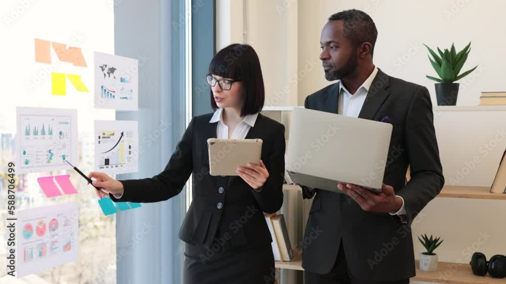 Young caucasian businesswoman with tablet in hand pointing at bar graphs while multicultural co-worker checking data on laptop. Professional partners analysing financial statistics using documents. Young caucasian businesswoman with tablet in hand pointing at bar graphs while multicultural co-worker checking data on laptop. Professional partners analysing financial statistics using documents.