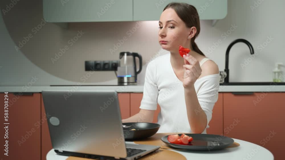 Young woman eats salad and pasta while working on a laptop, sitting at a table in the kitchen at home. A business woman in casual clothes has lunch while working from home Young woman eats salad and pasta while working on a laptop, sitting at a table in the kitchen at home. A business woman in casual clothes has lunch while working from home