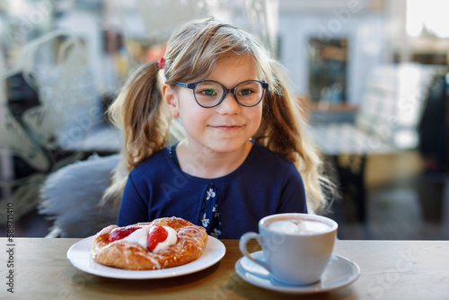 Adorable smiling girl with glasses have a breakfast in a cafe. Preschool child with glasses drinking chocolate and eating bakery pastry croissant or cake. Happy children, healthy food and meal.