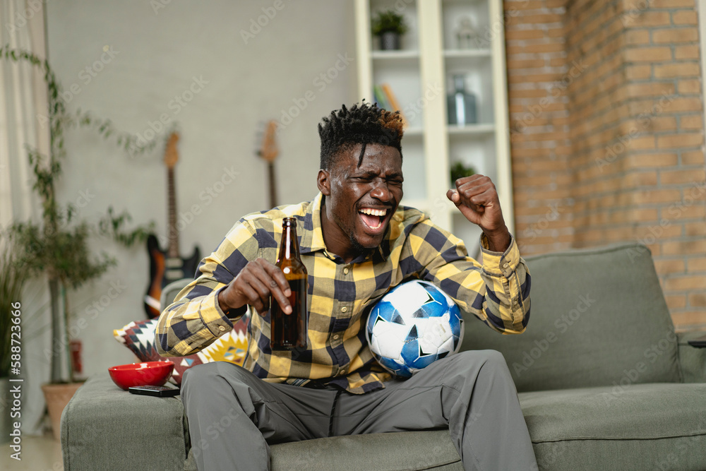Obraz premium Young African man watching sport match on television and supporting his favorite team. Happy man enjoying victory, watching sports competition on tv.