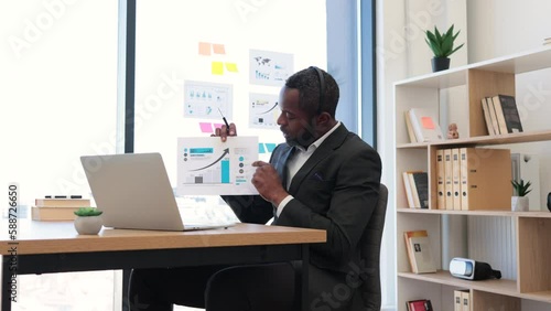 Formally dressed male entrepreneur sitting at workspace and having online meeting with colleagues on laptop. African american man in headset examining some documents holding in hands.