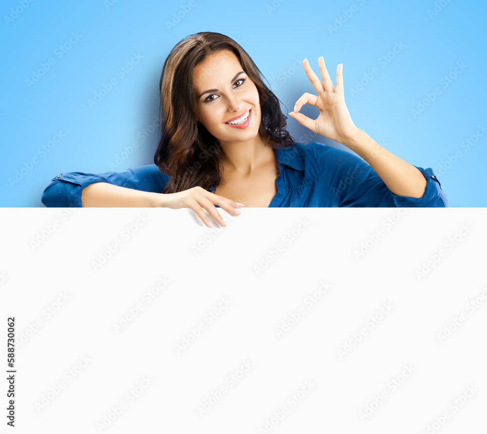 Happy smiling brunette young woman showing okay ok gesture, stand behind, peeping from blank banner or mock up signboard with copy space, isolated sky blue background. Happy smiling brunette young woman showing okay ok gesture, stand behind, peeping from blank banner or mock up signboard with copy space, isolated sky blue background.