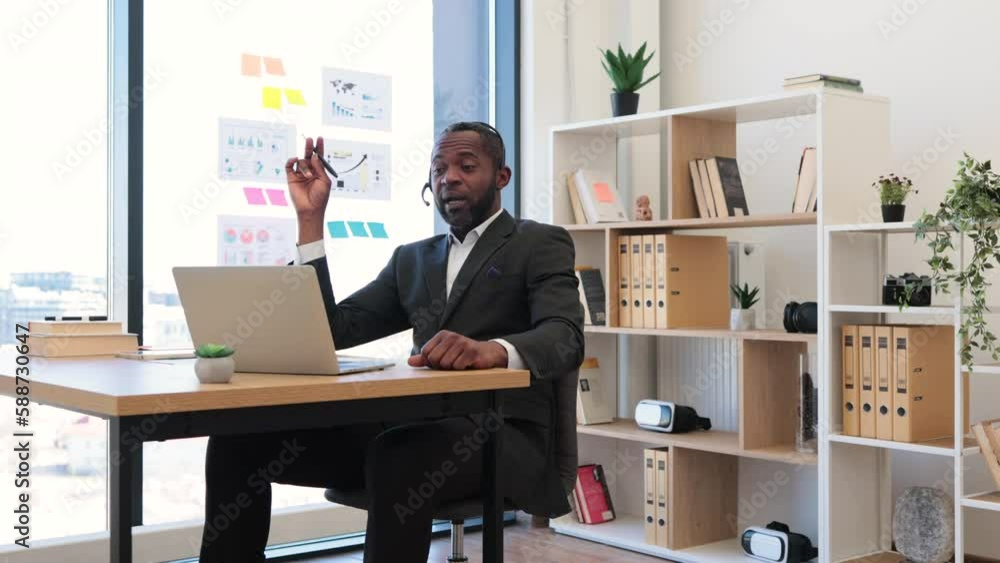 African american man in formal suit having video call at office using headset and wireless laptop. Transparent board with various graphs and charts on background. Concept of online business meeting. African american man in formal suit having video call at office using headset and wireless laptop. Transparent board with various graphs and charts on background. Concept of online business meeting.