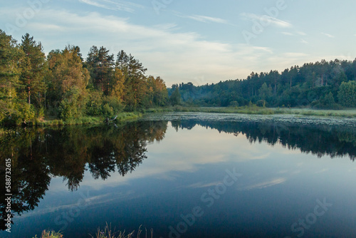 A picturesque reflection of the sky and trees in a forest river. The concept of vacation, summer, fishing, rest on the river bank, calendar, cover. Horizontal photo. 
