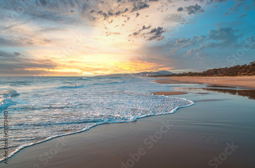 Mesmerizing sunset behind the clouds over pristine sandy beach on the Sunshine Coast in Australia. Wide angle beach panorama. 