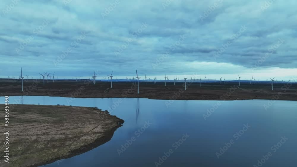 Aerial rising shot showing wind turbines spinning in a large windfarm next to a lake Aerial rising shot showing wind turbines spinning in a large windfarm next to a lake