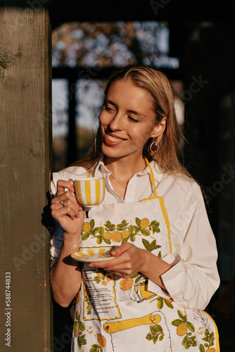 Cute smiling good-looking woman with wonderful smile wearing apron and white shirt is having coffee break while cooking outdoor in sunlight