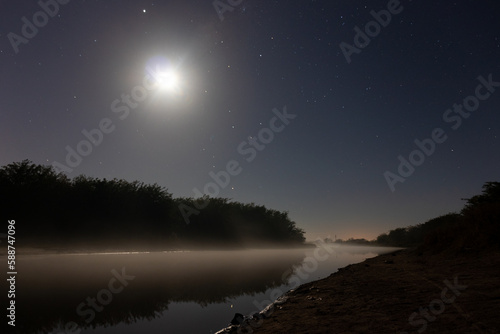 A lake at night, with calm waters and mist covering the surface. In the distance, shadowy trees are silhouetted against the mist and the moon and a few stars peek through the clouds. A magical and eni