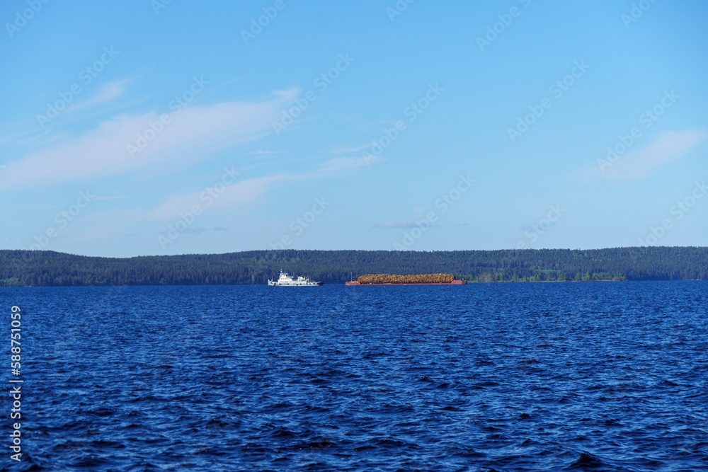 Tugboat pulls barge with cargo of commercial timber by lake water in summer