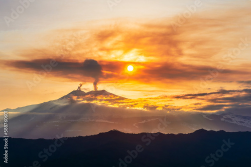 mysterious landscape of great erupting volcano with smoke from craters and snow on slopes in orange light of sunset. eruption of vulcan photo