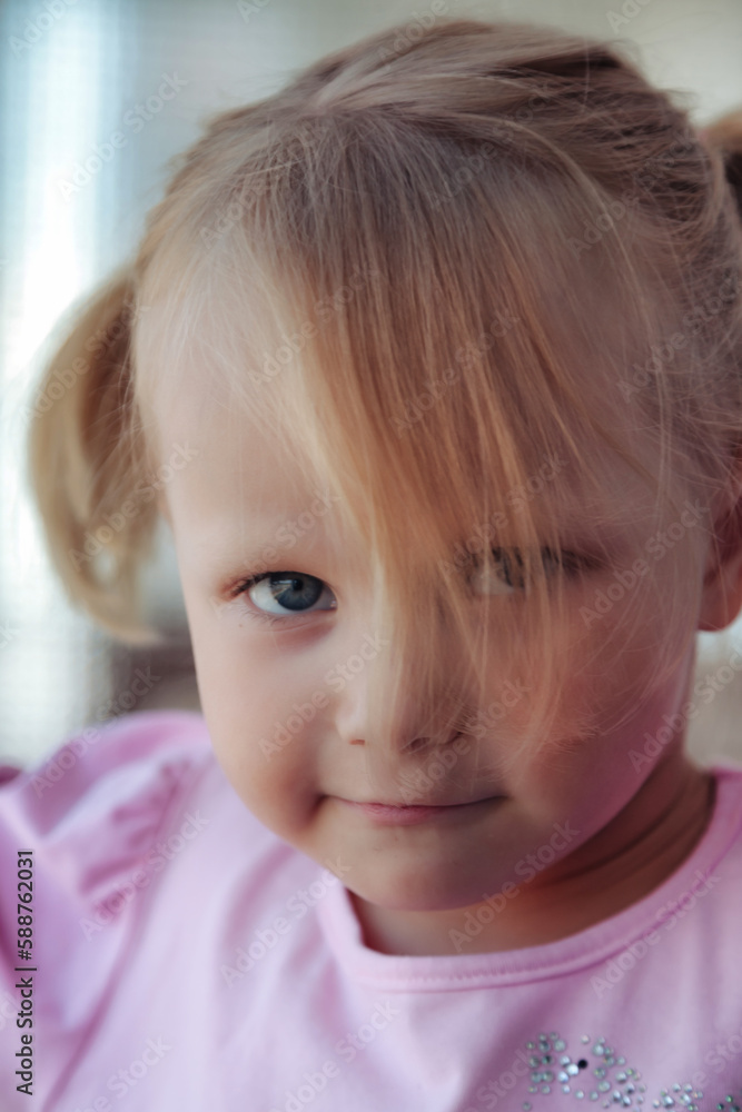 Face of smiling pensive little girl posing near window, thought looking at camera. Portrait adorable bored child with bangs on head indoors. Concept of childhood kids emotion. Copy ad text space
