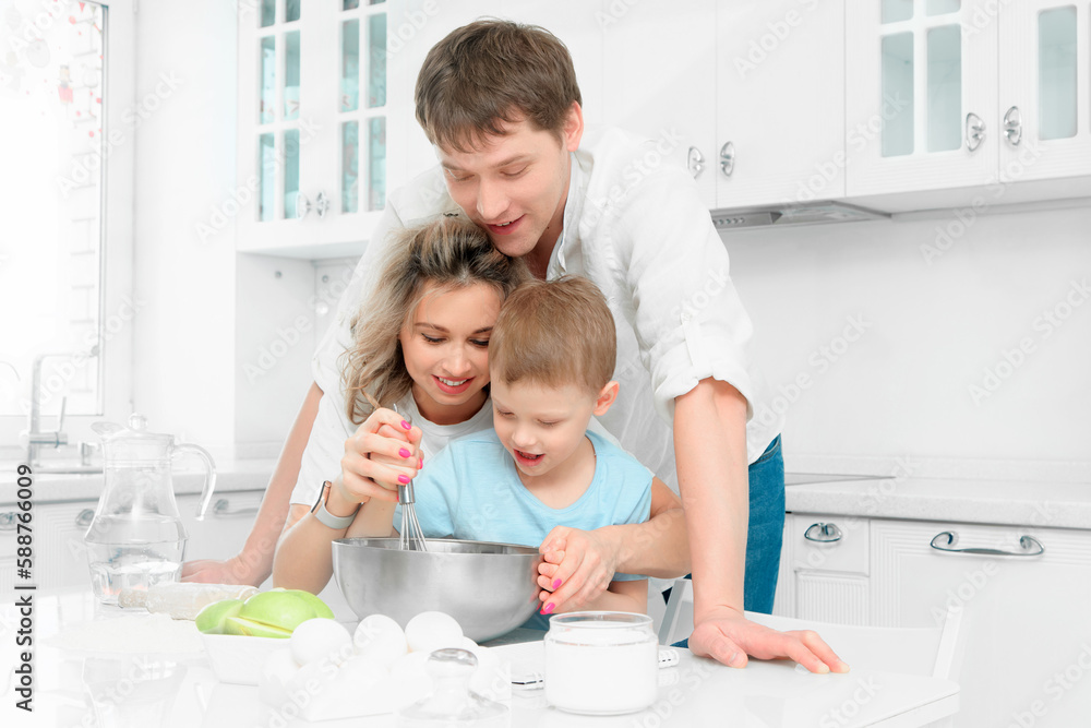 Young Caucasian family of three cooks lunch or festive cake in kitchen according to recipe. Happy family at table in bright kitchen.. Young Caucasian family of three cooks lunch or festive cake in kitchen according to recipe. Happy family at table in bright kitchen..