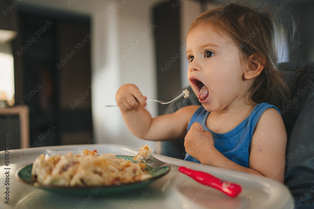 Toddler baby girl learnsing to eat herself with a fork while sitting in a child chair. Nutrition development. Toddler baby girl learnsing to eat herself with a fork while sitting in a child chair. Nutrition development.