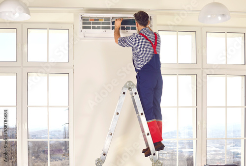 Professional electrician man standing back on a ladder maintaining or cleaning modern air conditioner indoors. Mature technician in the room repairing or installing air conditioner.
