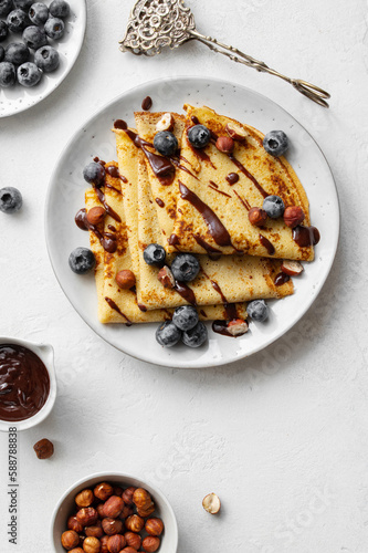 Crepes or blinis or thin pancakes with blueberries, hazelnuts and chocolate sauce on a white plate at white stone table. Maslenitsa traditional festival meal.