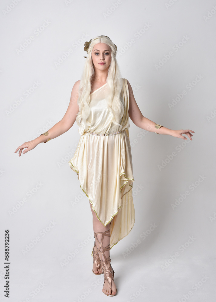 Full length portrait of beautiful blonde woman wearing a fantasy goddess toga costume with  magical crown.
Standing, dancing pose with gestural hands reaching out, isolated on white studio background.