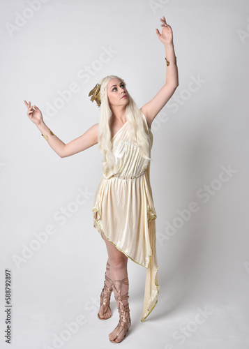 Full length portrait of beautiful blonde woman wearing a fantasy goddess toga costume with  magical crown.
Standing, dancing pose with gestural hands reaching out, isolated on white studio background.