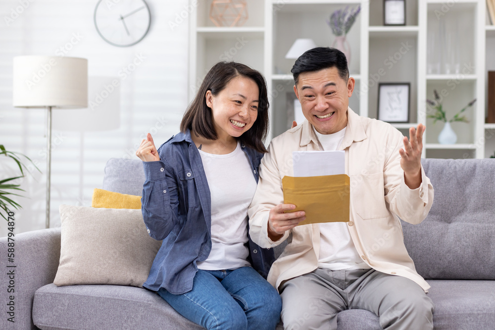 Cheerful and happy asian family together, man and woman at home on sofa sitting in living room, received postal envelope with good news, message and good achievement news. Cheerful and happy asian family together, man and woman at home on sofa sitting in living room, received postal envelope with good news, message and good achievement news.