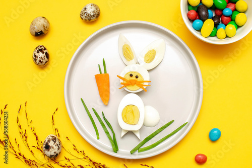 Easter funny creative healthy breakfast lunch food idea for kids, children.Bunny, rabbit made from boiled chicken eggs,peeled carrots, greens on plate yellow table background.Top view Flat lay