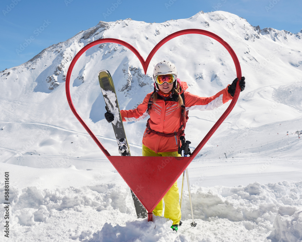 Woman skier poses inside a heart shaped frame at Tignes - Val d'Isere ski resort, France, with all mountan skis and red outfit, on a sunny day. Tourism, activity, Winter, snow.