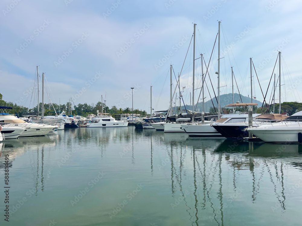 Luxury boats in marina. Yacht club. Sailboats on the harbour. Reflection of masts on a sea surface. Calm bay. Sky with clouds. Moored vessels. Tropical islands. Land on the horizon. Sailing, yachting