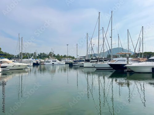 Luxury boats in marina. Yacht club. Sailboats on the harbour. Reflection of masts on a sea surface. Calm bay. Sky with clouds. Moored vessels. Tropical islands. Land on the horizon. Sailing, yachting