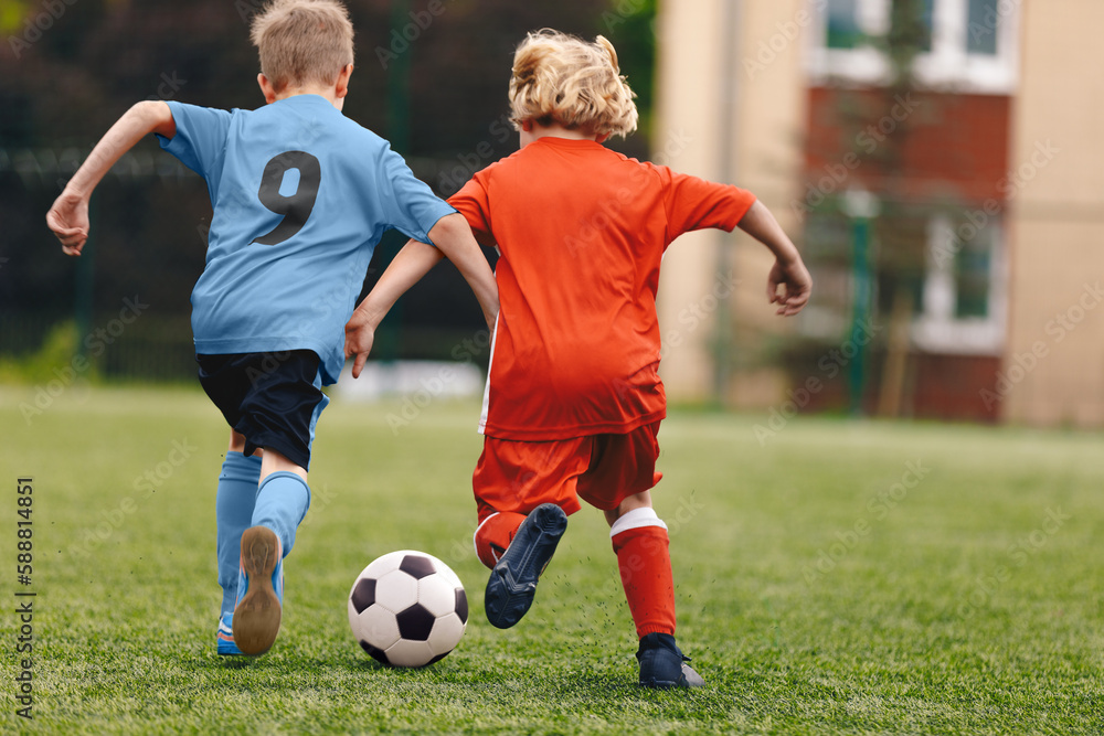 Two school boys kicking a soccer ball. School boys in red and blue football uniforms compete in a soccer match. Football duel in children soccer game Two school boys kicking a soccer ball. School boys in red and blue football uniforms compete in a soccer match. Football duel in children soccer game