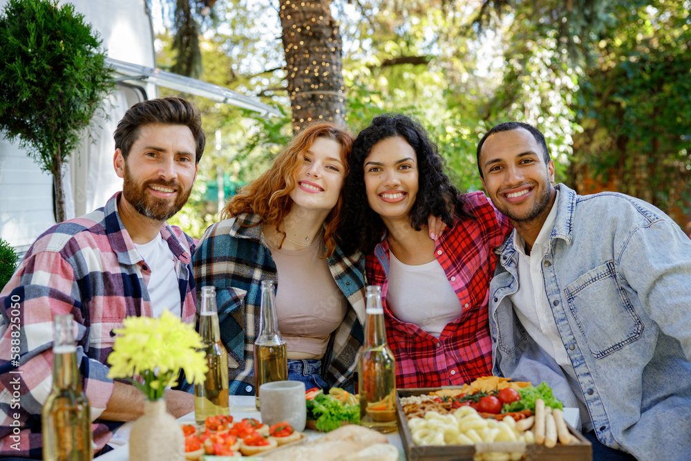 A group of friends are relaxing in nature. Two girls and two boys of different races are looking at the camera, smiling, having fun on the background of a travel trailer. A group of friends are relaxing in nature. Two girls and two boys of different races are looking at the camera, smiling, having fun on the background of a travel trailer.