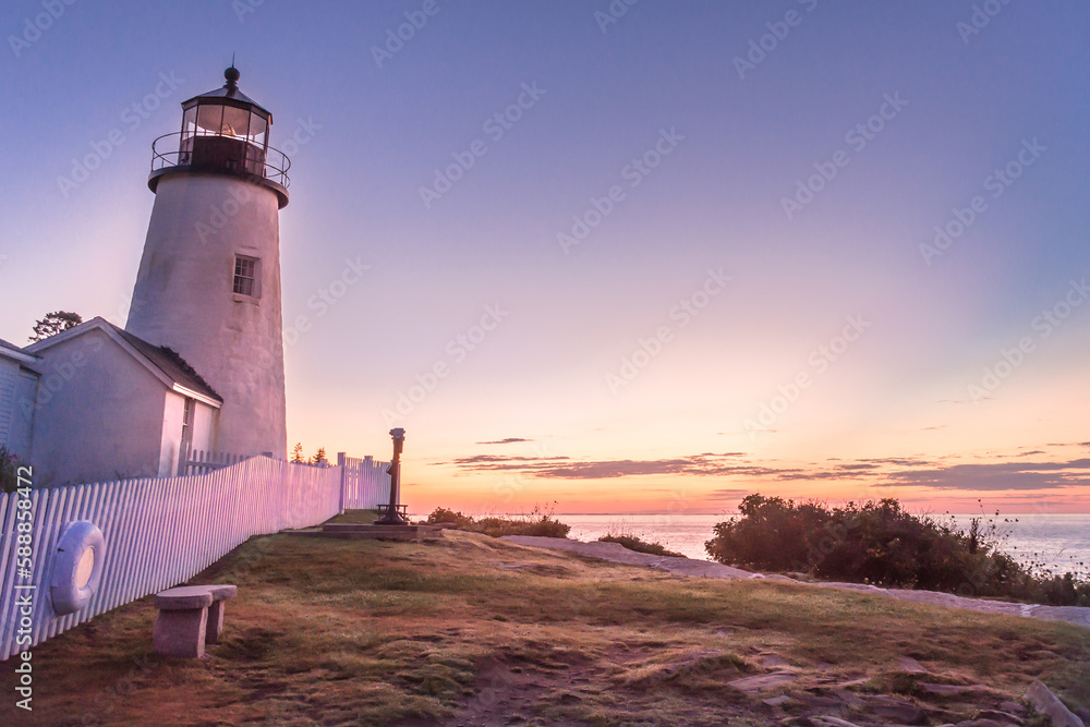 Morning sunrise at Pemaquid Lighthouse. Lighthouse and keeper's house bathed in rosy morning sun. Picket fence leading towards horizon. Fiery clouds on horizon over ocean. Deep blue morning sky.