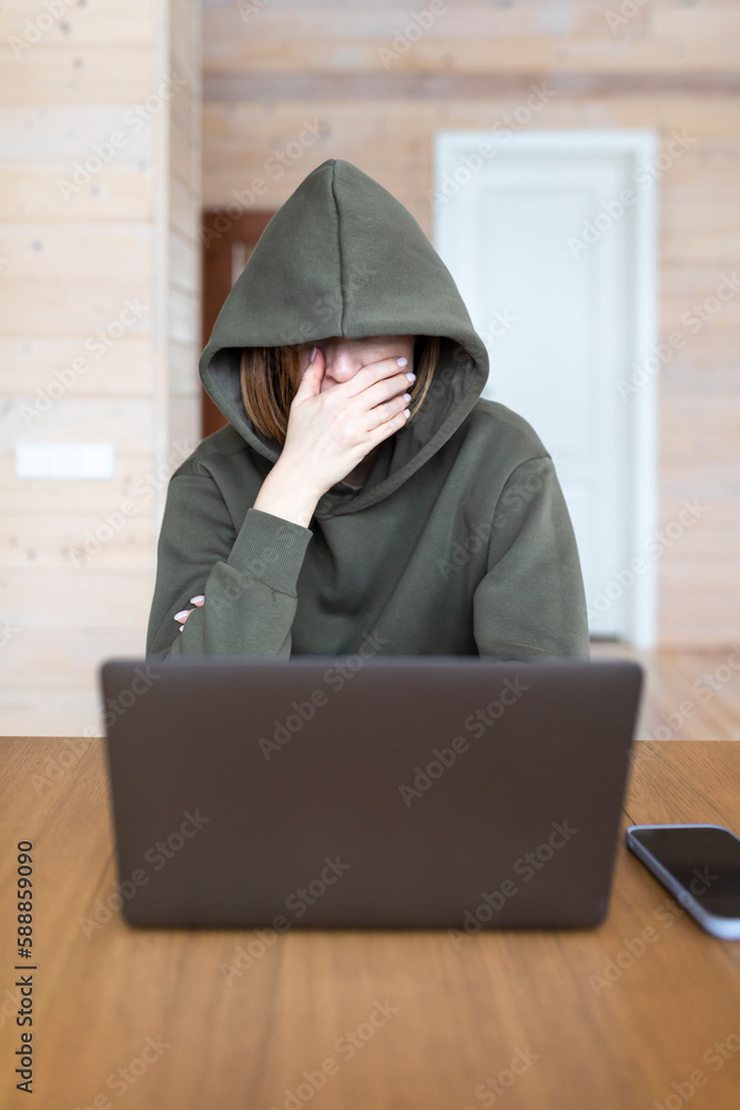 Portrait of enigmatic woman in hoodie sits at the desk and works on laptop Portrait of enigmatic woman in hoodie sits at the desk and works on laptop