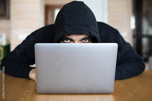 Canvas Print Portrait of enigmatic man in hoodie sits at the desk and works on laptop