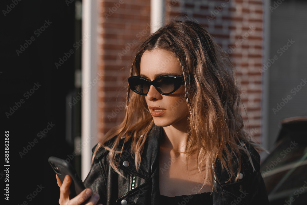 Outdoor shot of serious woman uses modern gadgets walks at street uses mobile phone, wears black glasses, leather jacket and dress sends sms tries to find route in unknown place in the city. Outdoor shot of serious woman uses modern gadgets walks at street uses mobile phone, wears black glasses, leather jacket and dress sends sms tries to find route in unknown place in the city.